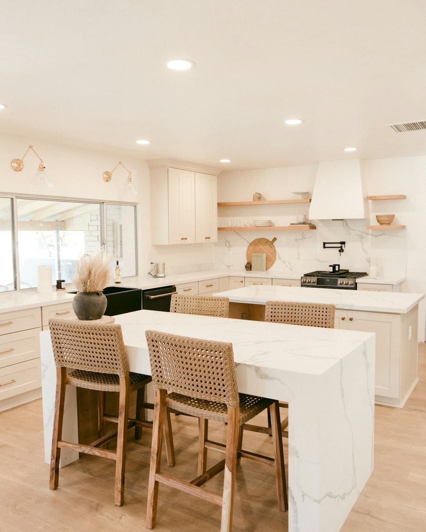 A warm, sunlit kitchen with soft neutral cabinetry, open wood shelving and a marble-topped island set for everyday living.