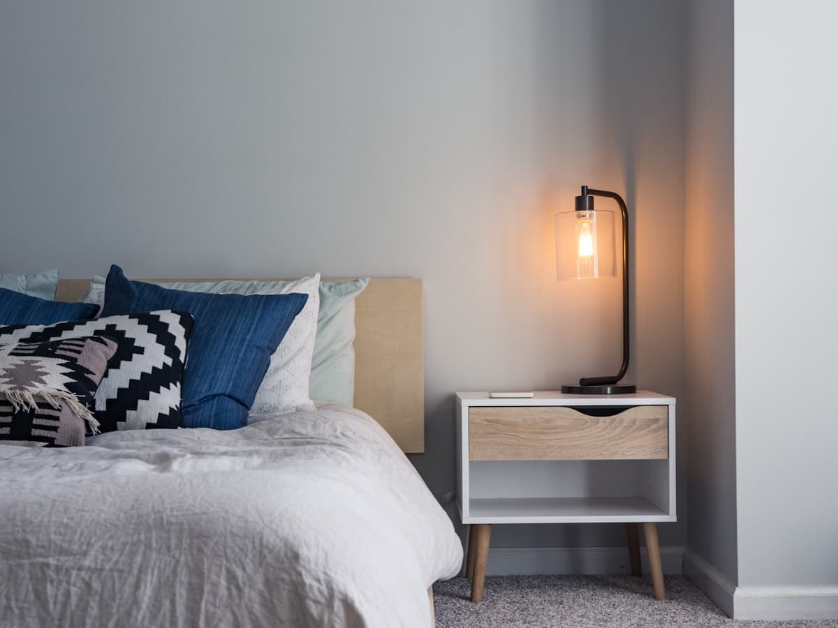 A warm bedside lamp casting soft light beside a bed in the evening.
